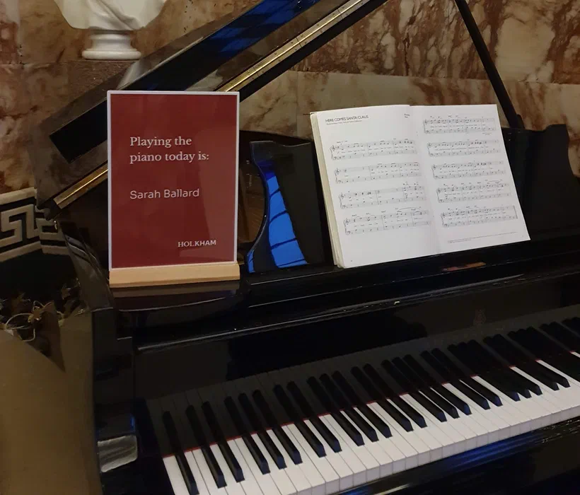 Grand piano at Holkham Hall with a sign reading 'Playing the piano today is: Sarah Ballard', keyboard and sheet music in view