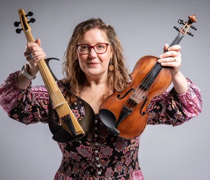 Sarah holding up both her violins to the camera — her Yamaha electric in one hand, her 300-year-old acoustic in the other
